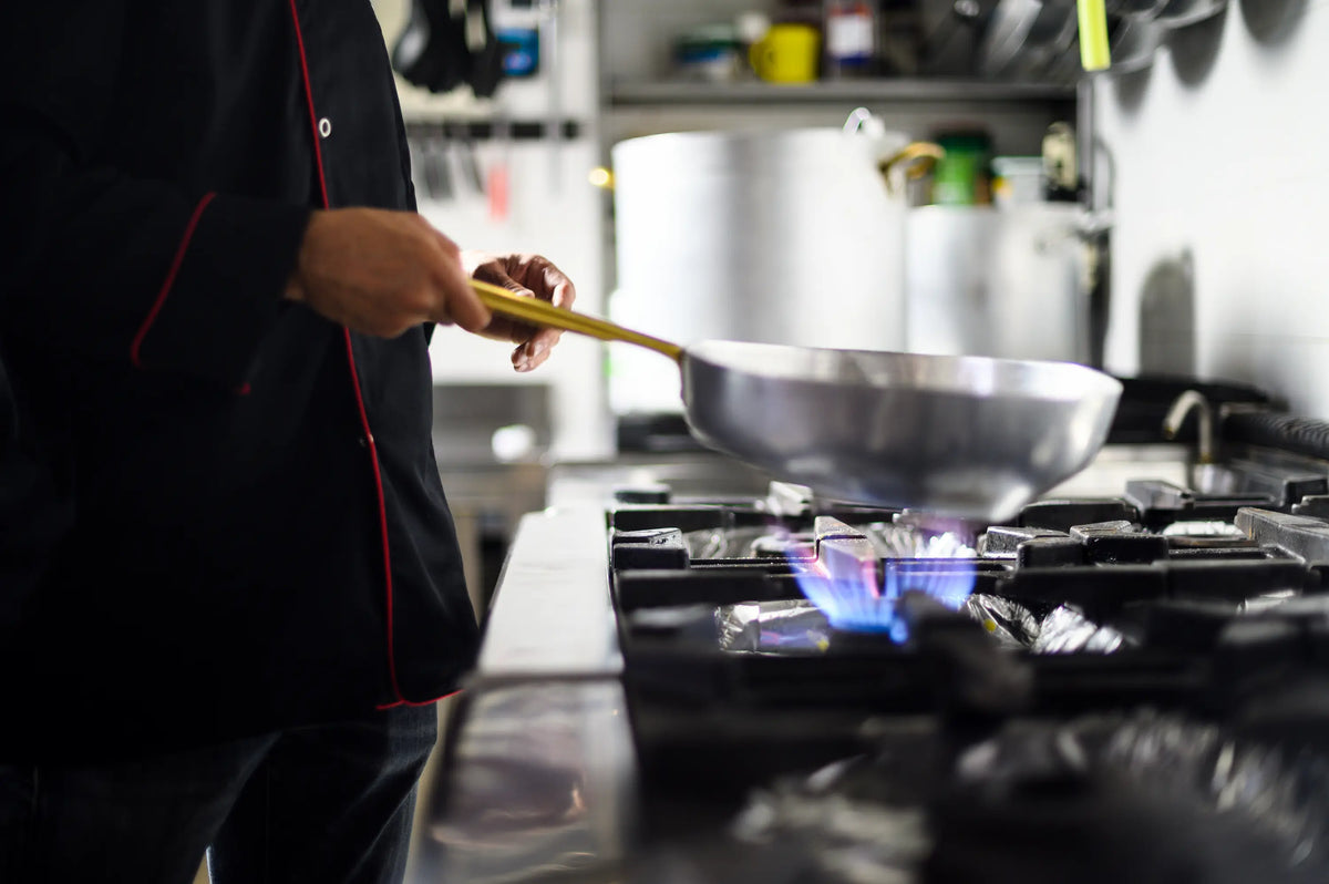 chef cuisinier avec une poele en inox dans les mains entrain de faire a manger dans une cuisine d'un restaurant avec un gazinière