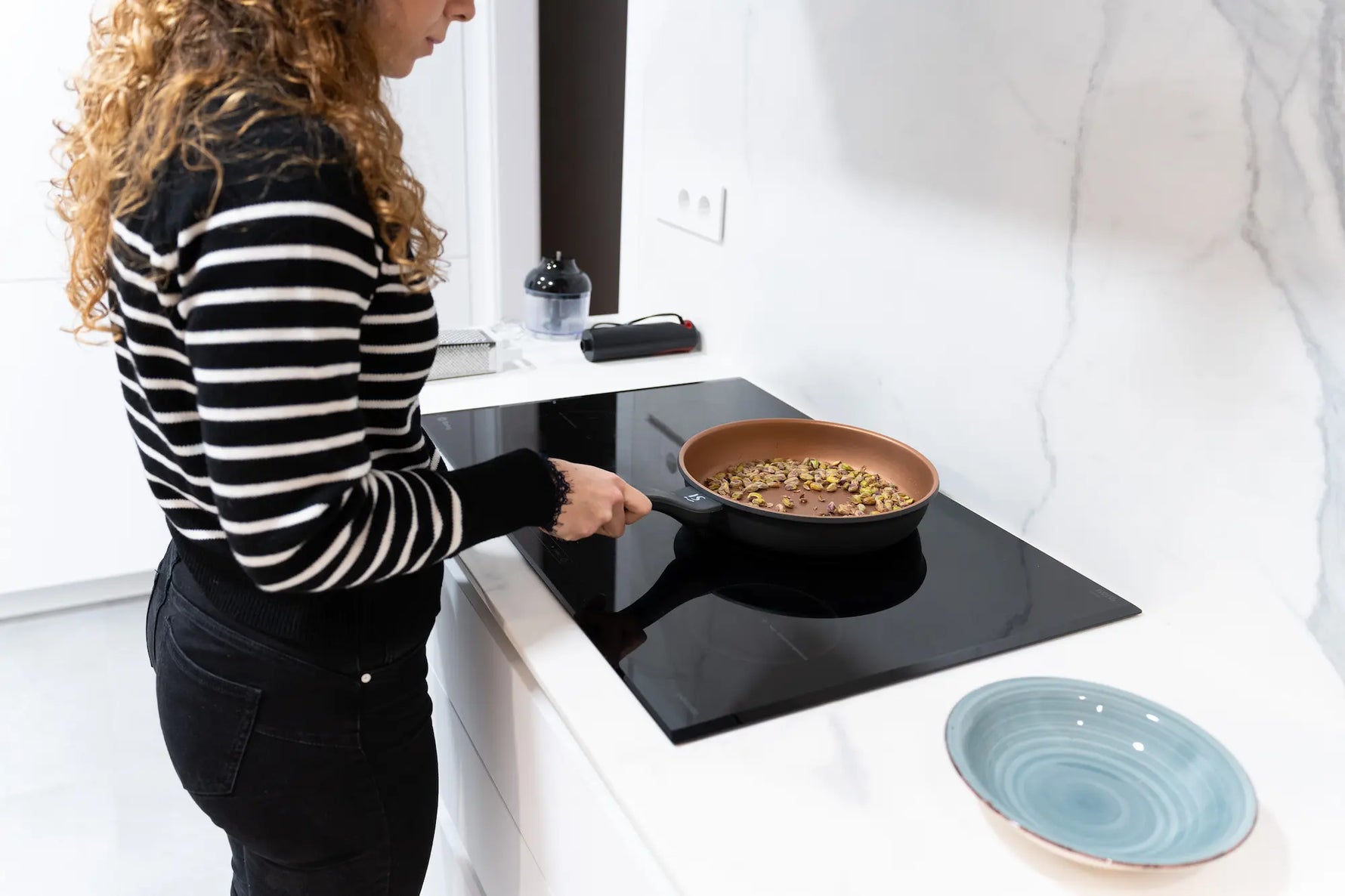 femme au cheveux bouclé habillé tout en noir entrain de faire torréfier des pistaches dans sa poele en ceramique sur une plaque à induction 
