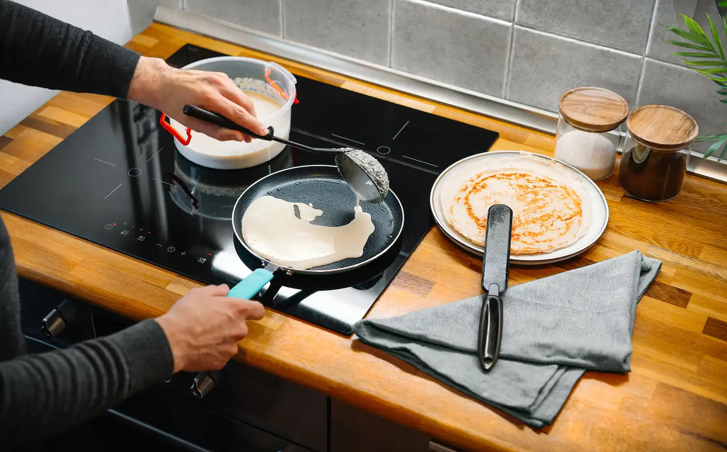 homme qui etale sa pate à crepe dans une poele avec un manche bleu sur une plaque a induction sur un plan de travail en bois avec une assiette et un torchon dessus
