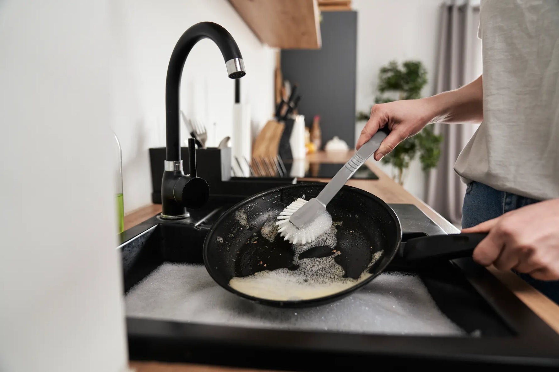 homme  qui nettoie une poele avec une brosse grise et du savon dans son evier de cuisine