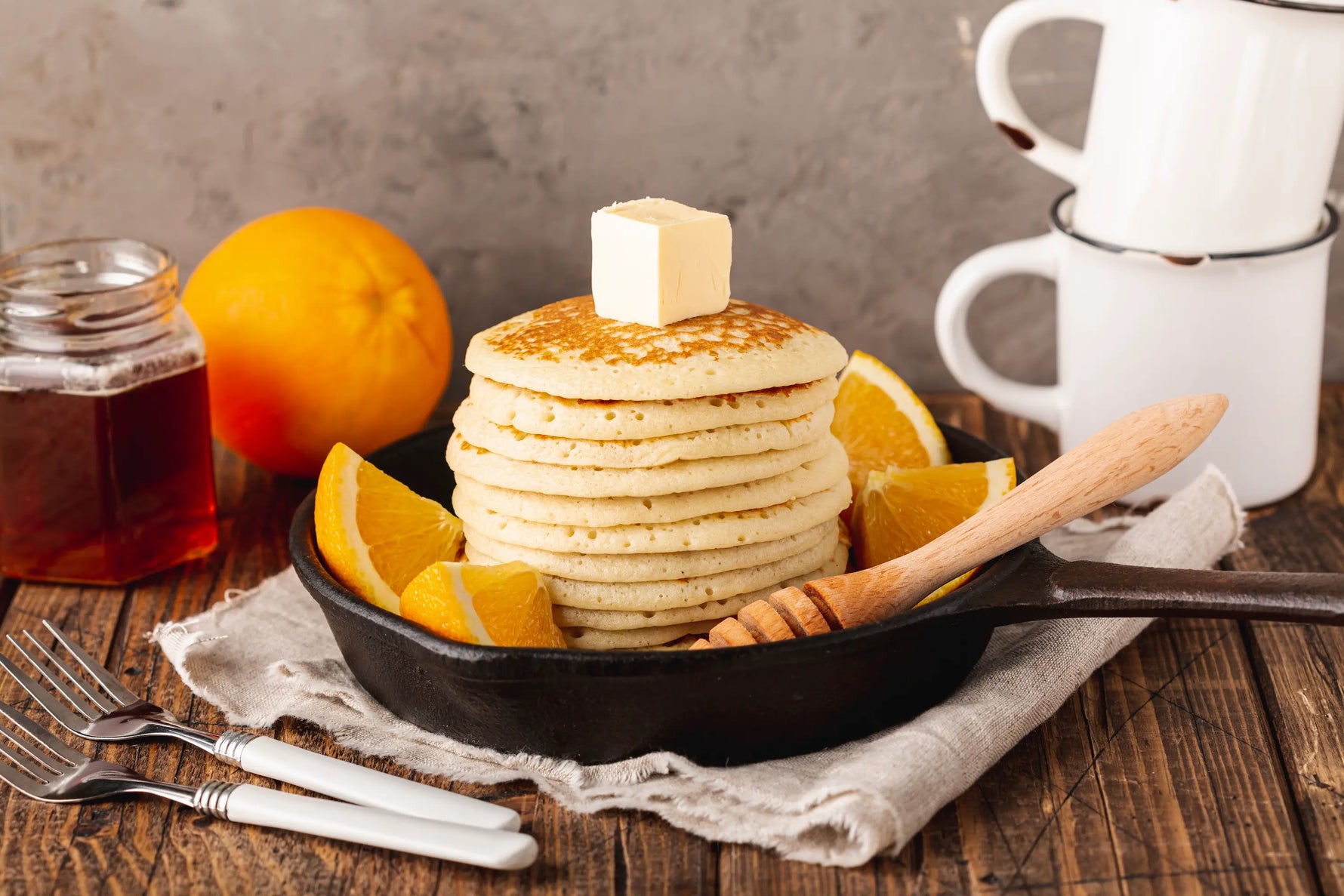 pancakes parfait dans une poele avec des quartiers d'oranges sur une table avec deux fourchettes et tasses accompagné de miel 