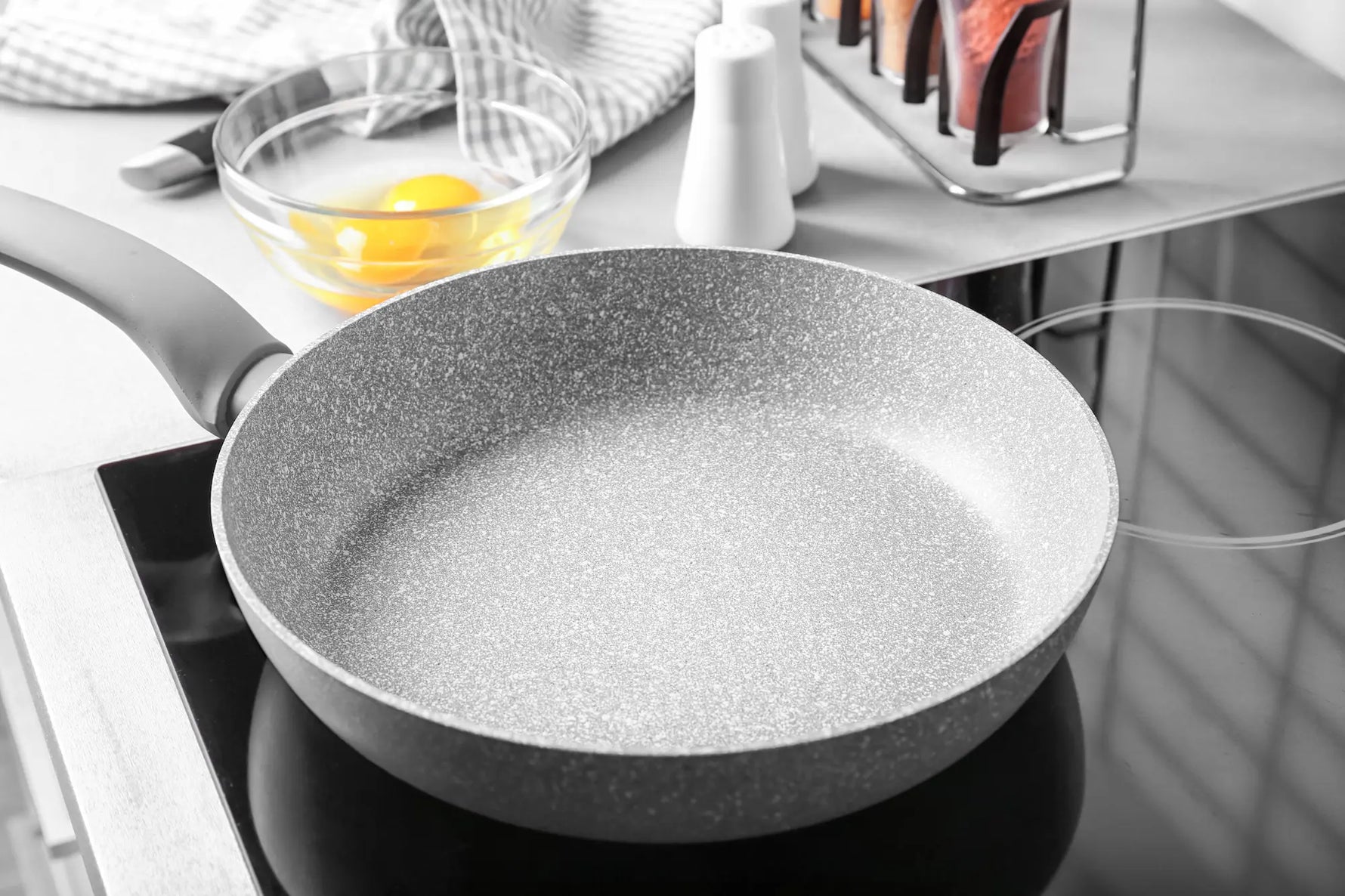 poele en pierre gris claire sur une plaque a induction avec en arriere plan un torchon un couteau de cuisine et des oeufs dans un bol transparent 
