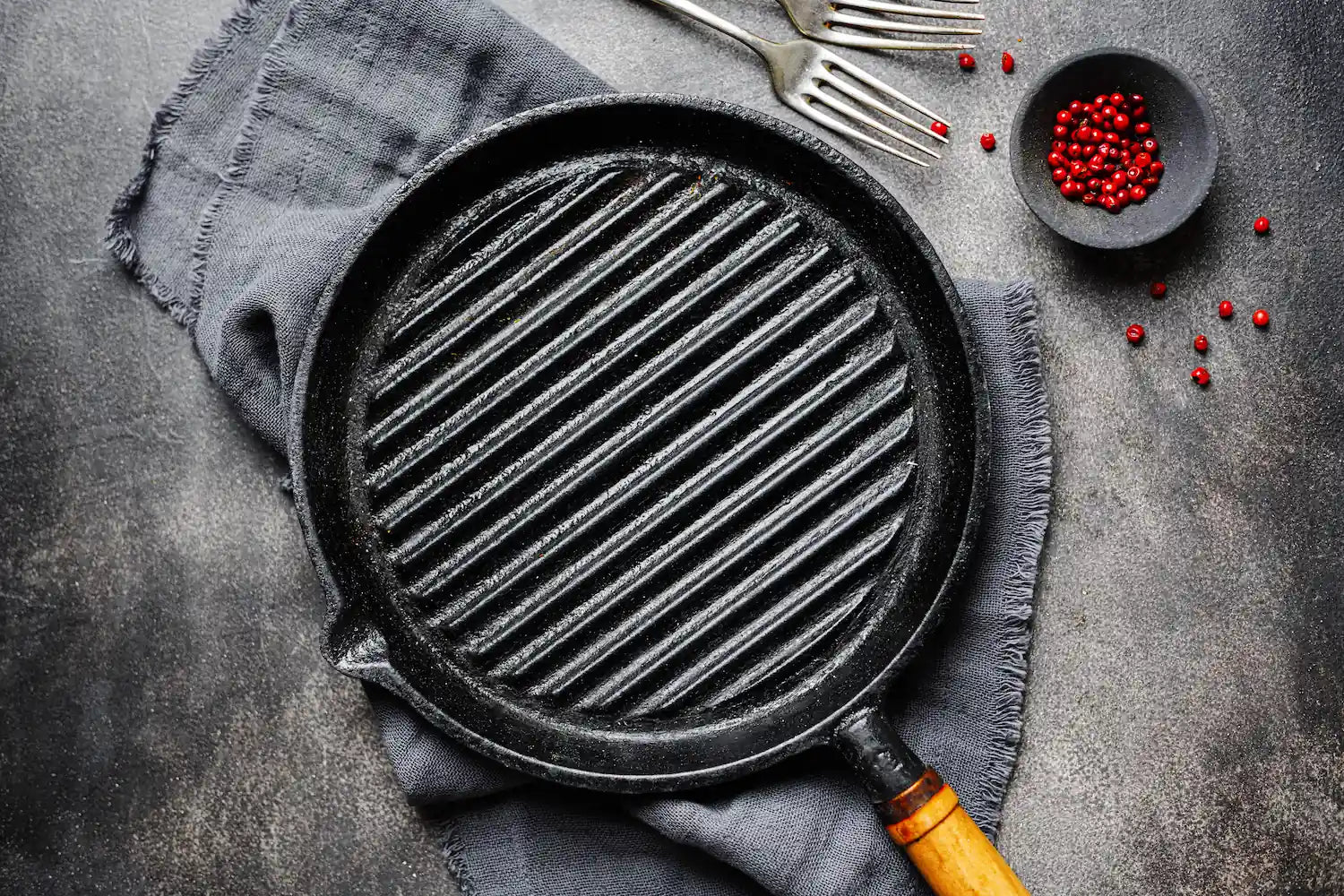 poele à grill avec un manche en bois sur un torchon sur une table dressé  avec des fourchettes et du poivres rouges