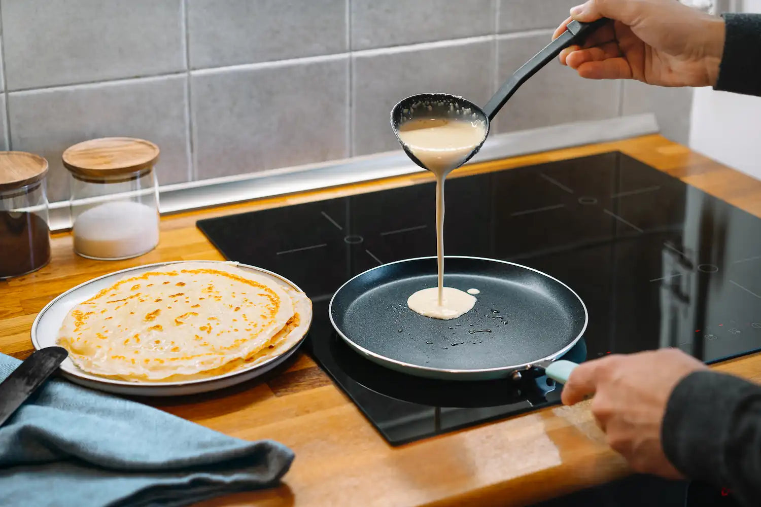 homme qui prepare des crepe dans sa poele à induction avec une assiette rempli de crepe deja cuite 