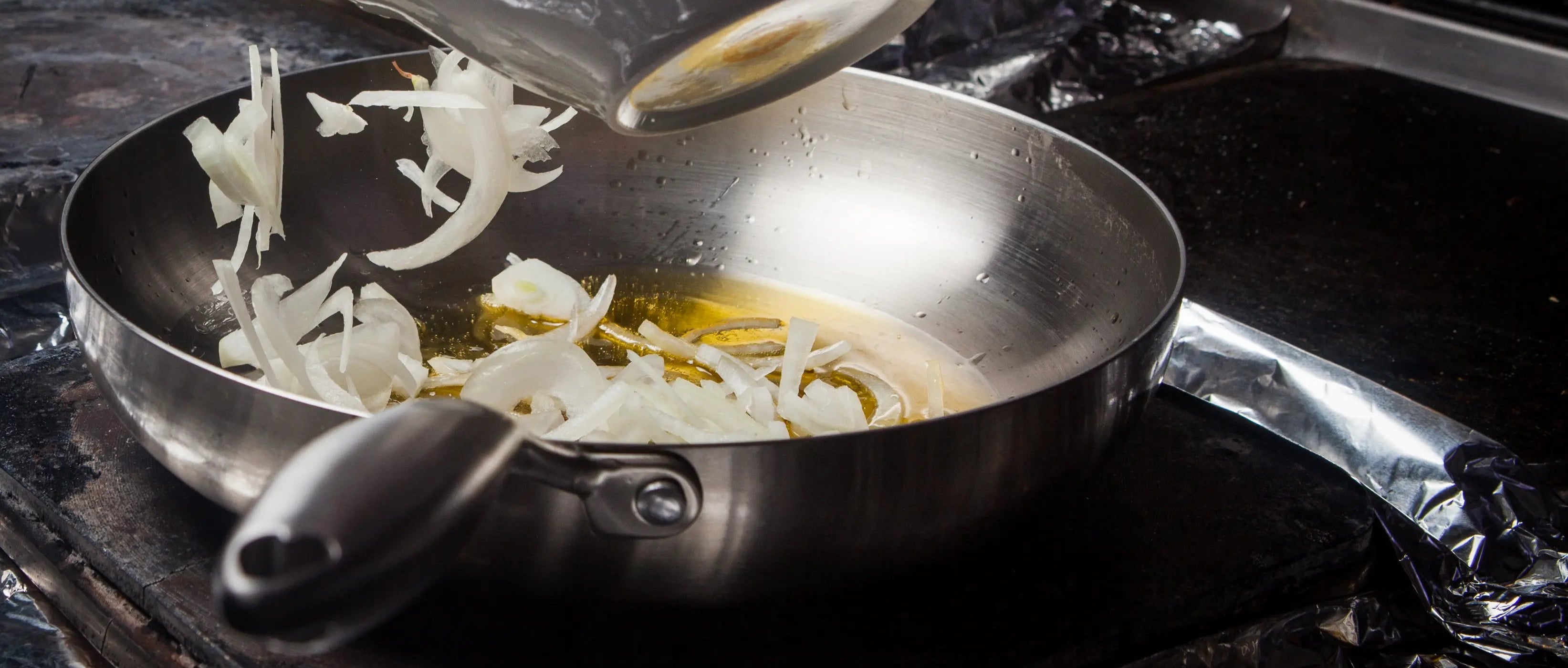 bol qui renverse des oignons coupés en lamelle dans une poêle en inox avec en arrière plan une cuisine de chef